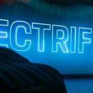 Neon blue sign of the word "ELECTRIFIED" behind the silhouette of two display vehicles